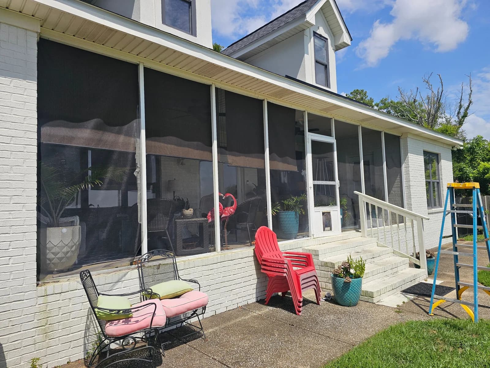 A two-story white-painted brick house with a covered front porch enclosed by dark roll-down shades and white metal porch posts, with an aluminum extension ladder leaning against the driveway-side column.