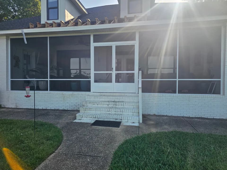 A two-story white-painted brick house with a new white aluminum-framed screened porch enclosure and double glass-paneled screen doors, with wood roof framing visible above the porch roofline, photographed against low sun with lens flare.