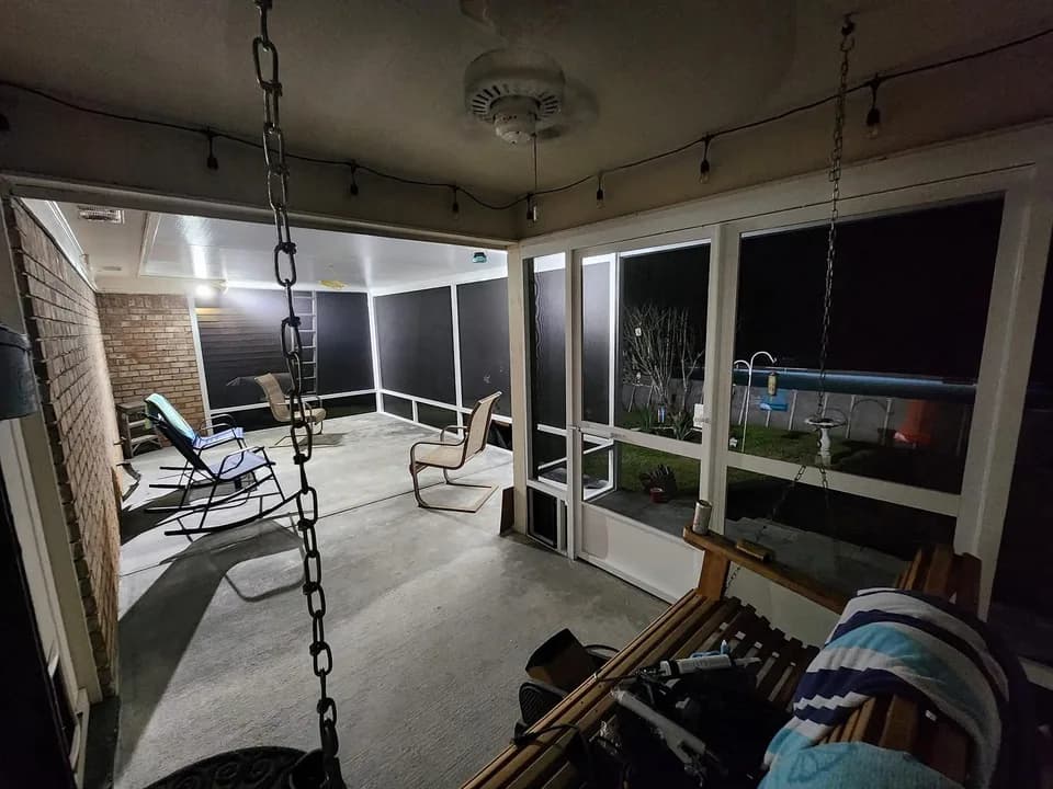 Interior night view of a screened back porch with a concrete floor, ceiling fan, and dark aluminum-framed screen panels, furnished with folding chairs and a porch swing, lit by string lights along the ceiling.