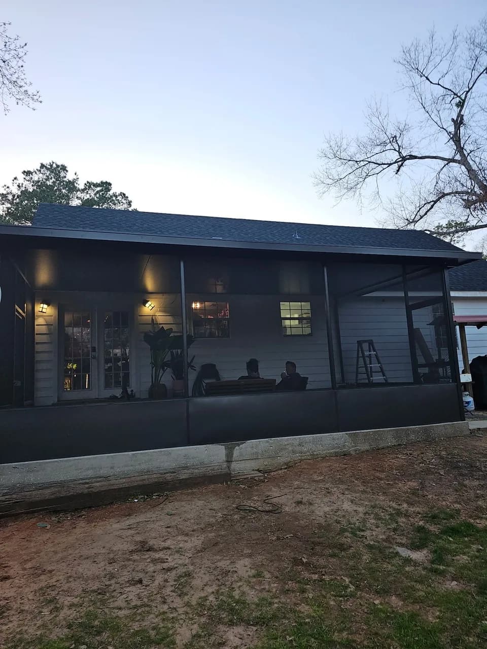 A dusk exterior view of a house with a dark bronze-framed aluminum screened porch addition, interior lights on, showing a stepladder and two people seated inside near a table.