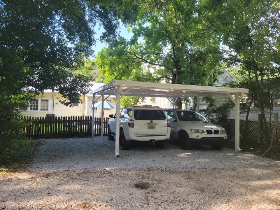 A freestanding white aluminum carport with a flat roof on four posts, sheltering two SUVs parked side by side on a gravel driveway, next to a single-story house with a wood picket fence, under shade trees.