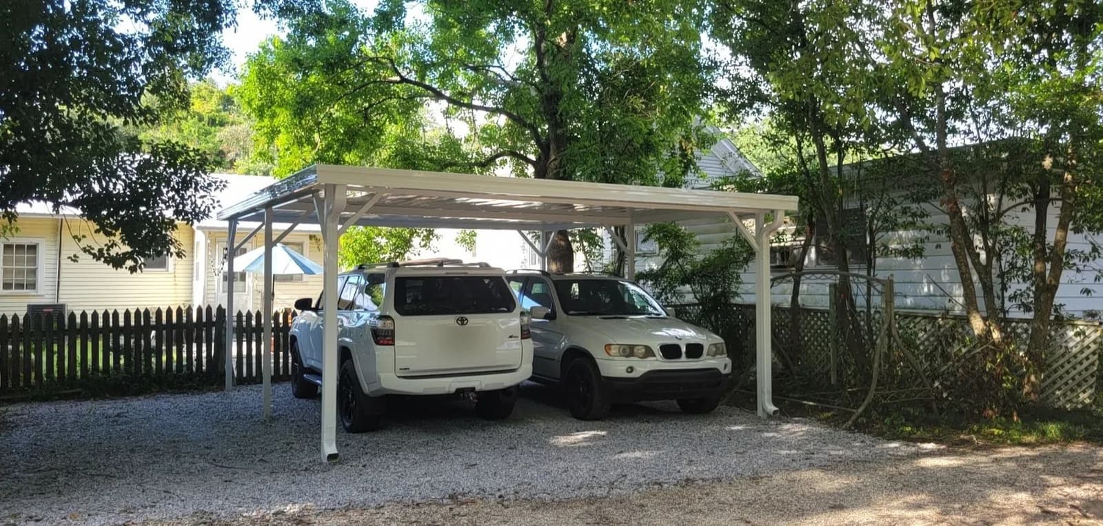 A freestanding white aluminum carport sheltering two SUVs on a gravel driveway next to a picket fence and a pale-yellow house, viewed from the front corner.