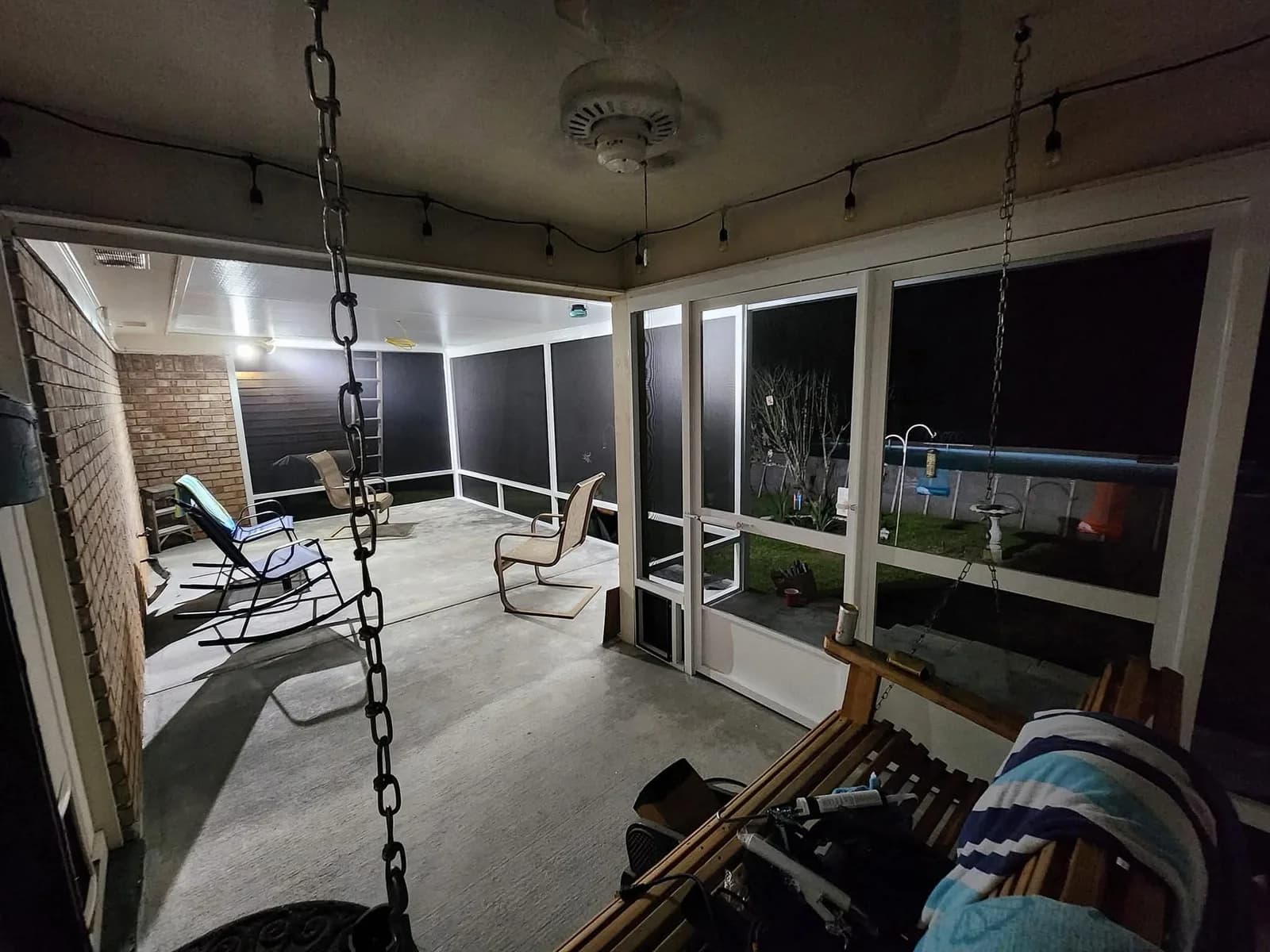Interior night view of a screened back porch with a concrete floor, ceiling fan, and dark aluminum-framed screen panels, furnished with folding chairs and a porch swing, lit by string lights along the ceiling.