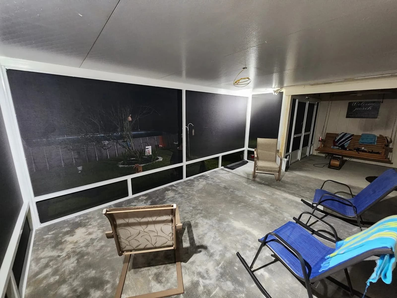 Interior night view of a screened porch corner with a hanging porch swing, patio lounge chairs, and a lit sign reading Welcome Porch mounted on the exterior wall, enclosed by dark aluminum-framed screen panels.