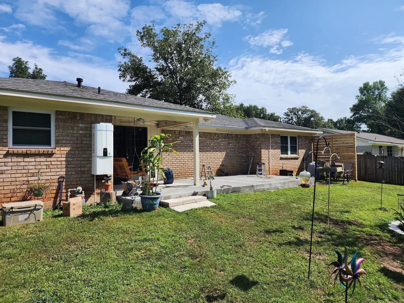 A brick ranch house with a newly built white aluminum patio roof cover over a bare gray concrete slab, open on all sides, with two stepladders on the slab and a horizontal wood-slat privacy screen at one end of the patio.